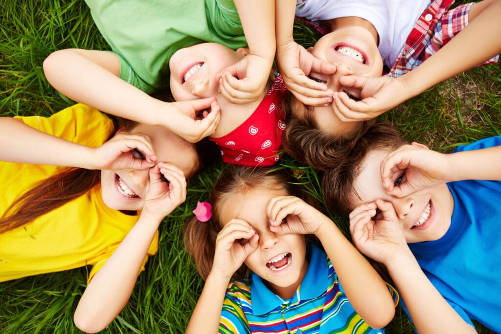 Group of joyful children lying on the grass, playing games with their hands, reminiscent of the carefree childhood fun described in the blog, where simple moments and imaginative games like paper planes and Chidiya Udd brought endless smiles.